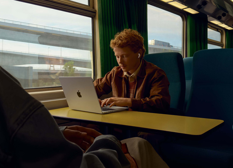 En person sidder ved en vinduesplads i et tog og arbejder på MacBook med AirPods i ørerne.