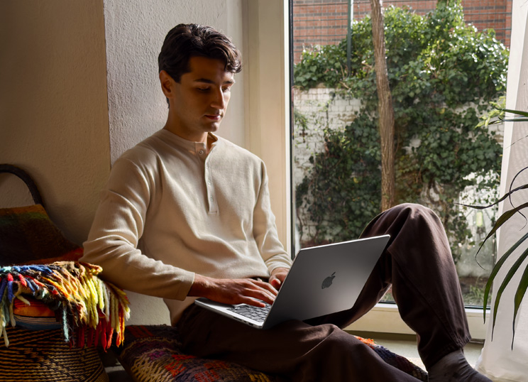 En person sidder på gulvet ved et vindue og bruger en MacBook.